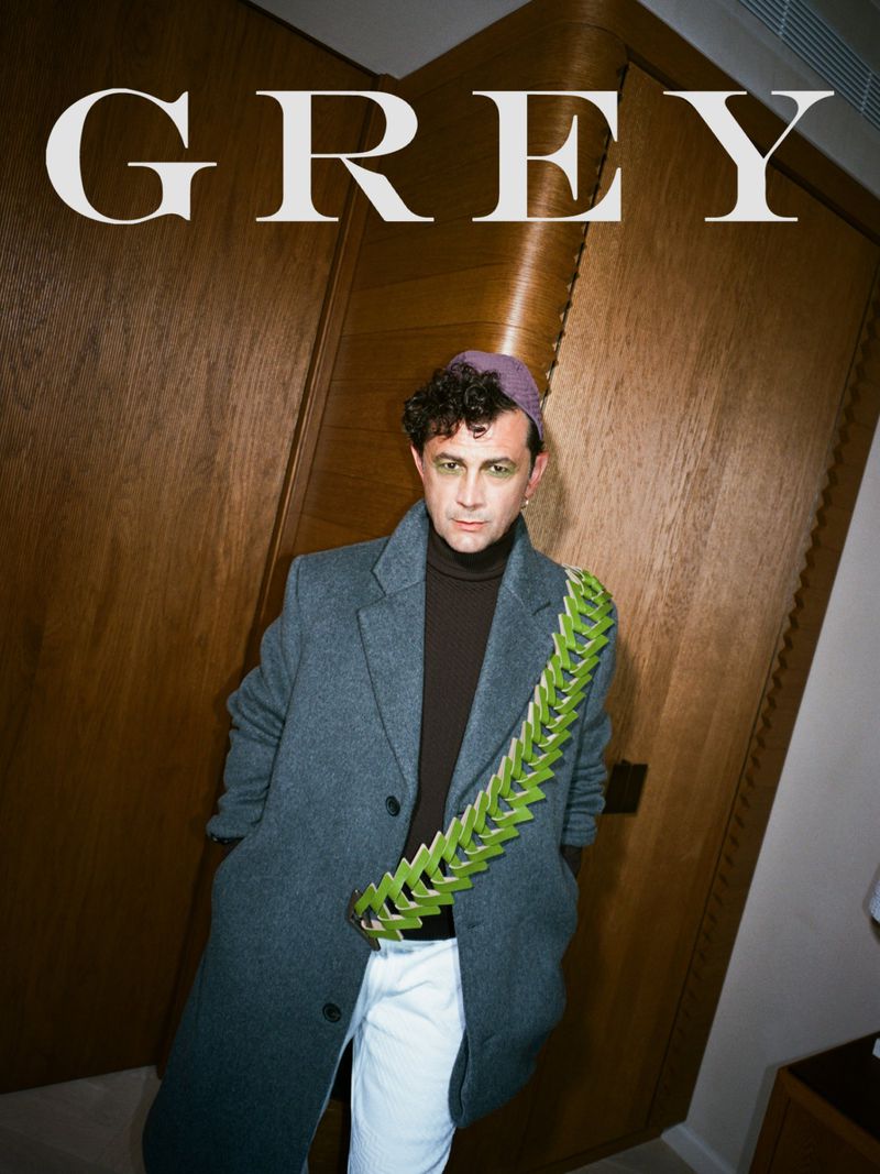 Magazine cover shot of a man wearing a grey coat and lilac cap standing against a wooden door with the text "GREY" at the top.