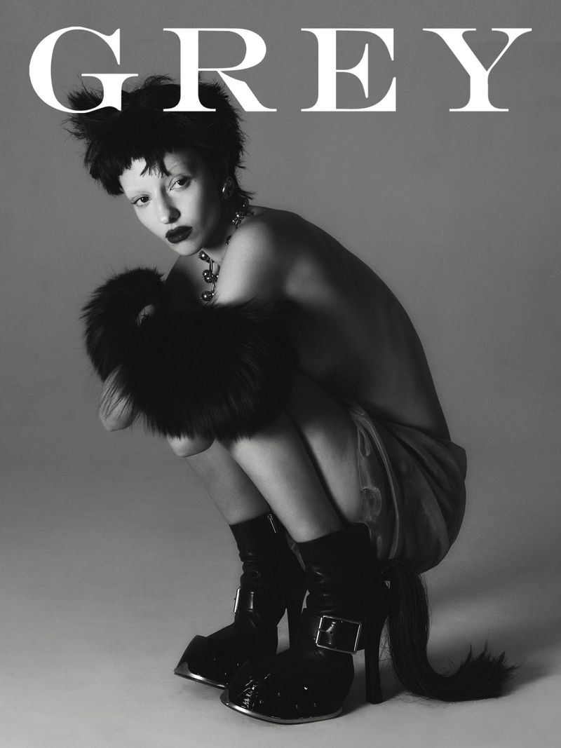 Black and white fashion shot of a crouching woman in a fur stole and boots with the word GREY in large white letters above her.