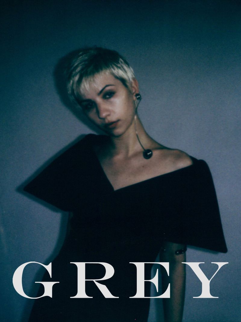 A woman with short blonde hair poses indoors wearing a black dress and long earring. The word 'GREY' is overlaid in white text.