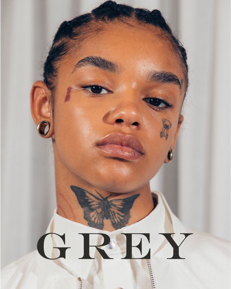 Close-up of a dark-skinned woman with braided hair, face tattoos, and ear tunnels, posing against a neutral backdrop. Fashion and beauty portrait.