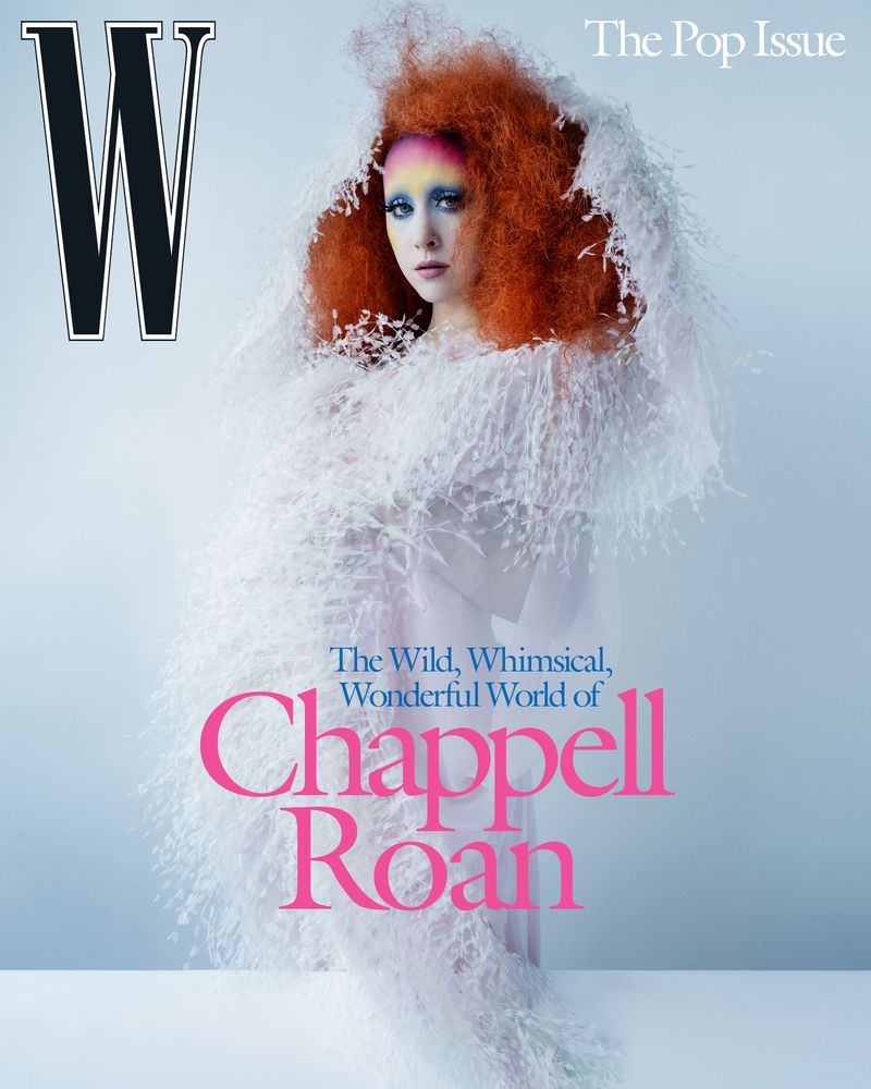 A person with voluminous red hair and vibrant makeup wears a white feathery garment against a light blue background.