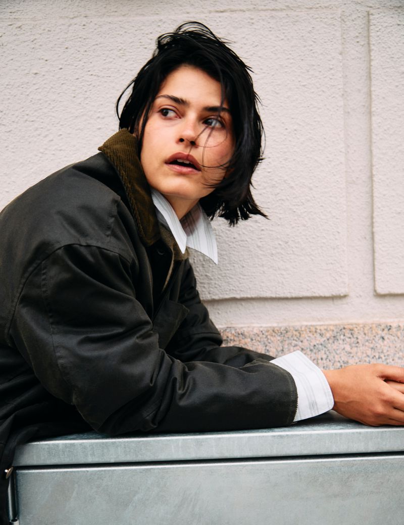 Brunette woman in jacket leans against a metal structure outdoors, looking to the side with a serious expression, wearing a white shirt.