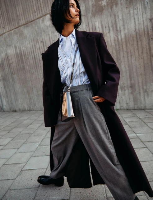 Elegant woman in oversized coat, light blue striped shirt, and gray trousers walking on paved surface in urban setting with concrete wall.