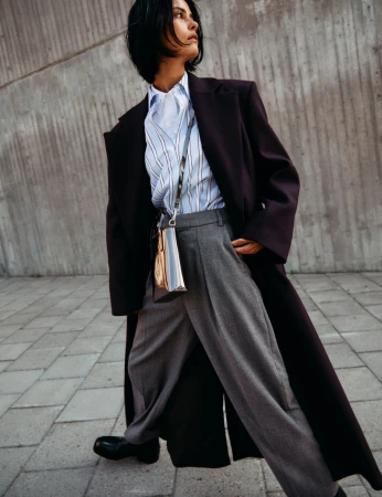Elegant woman in oversized coat, light blue striped shirt, and gray trousers walking on paved surface in urban setting with concrete wall.