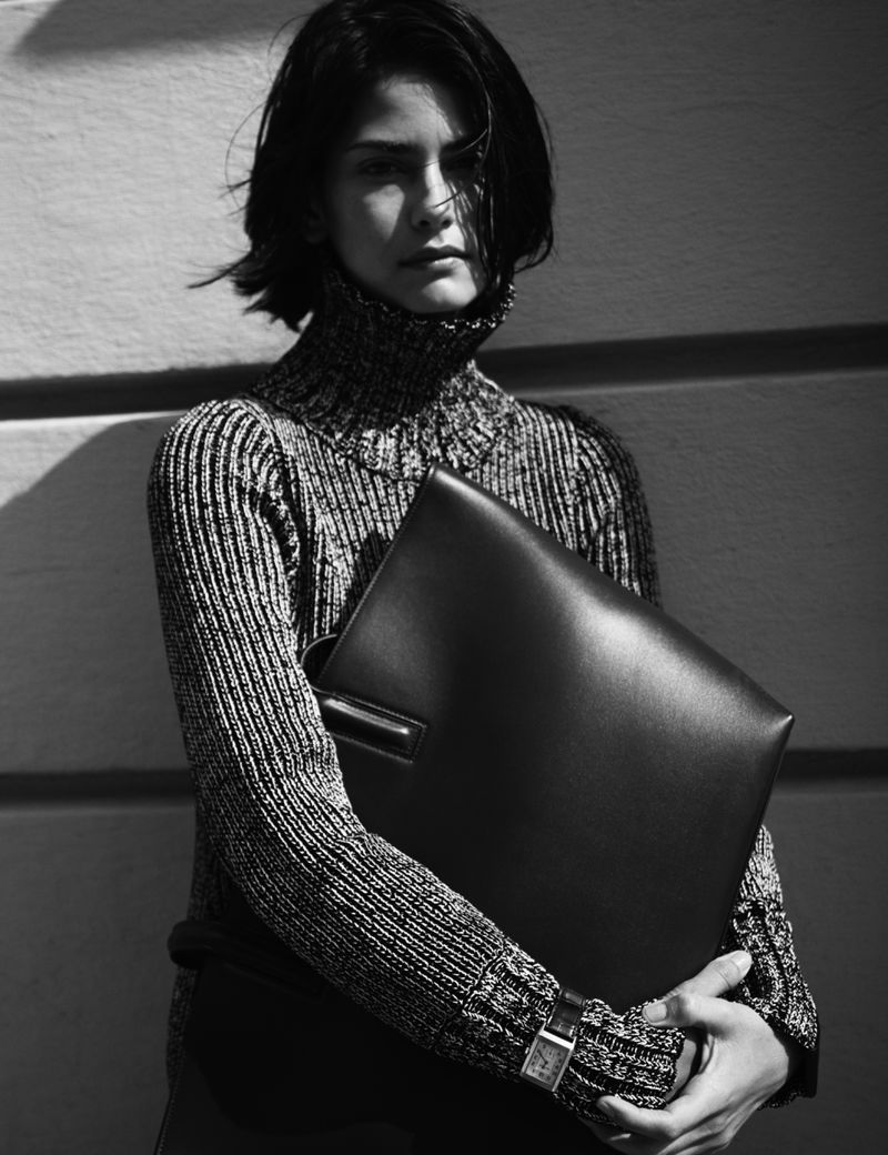 Model Hannah Kleit in a textured turtleneck sweater holding a large leather portfolio against a wall.