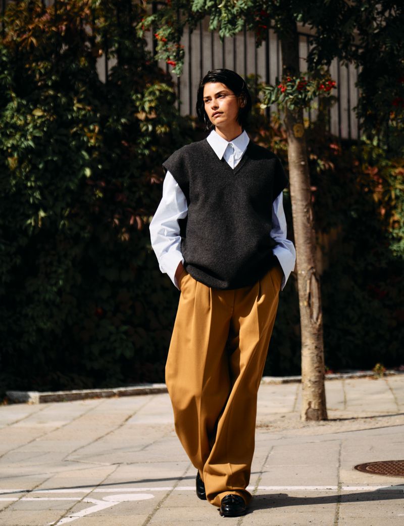 Hannah Kleit walking along a sidewalk wearing a dark knit sweater vest, white collared shirt, and wide-leg ochre trousers.