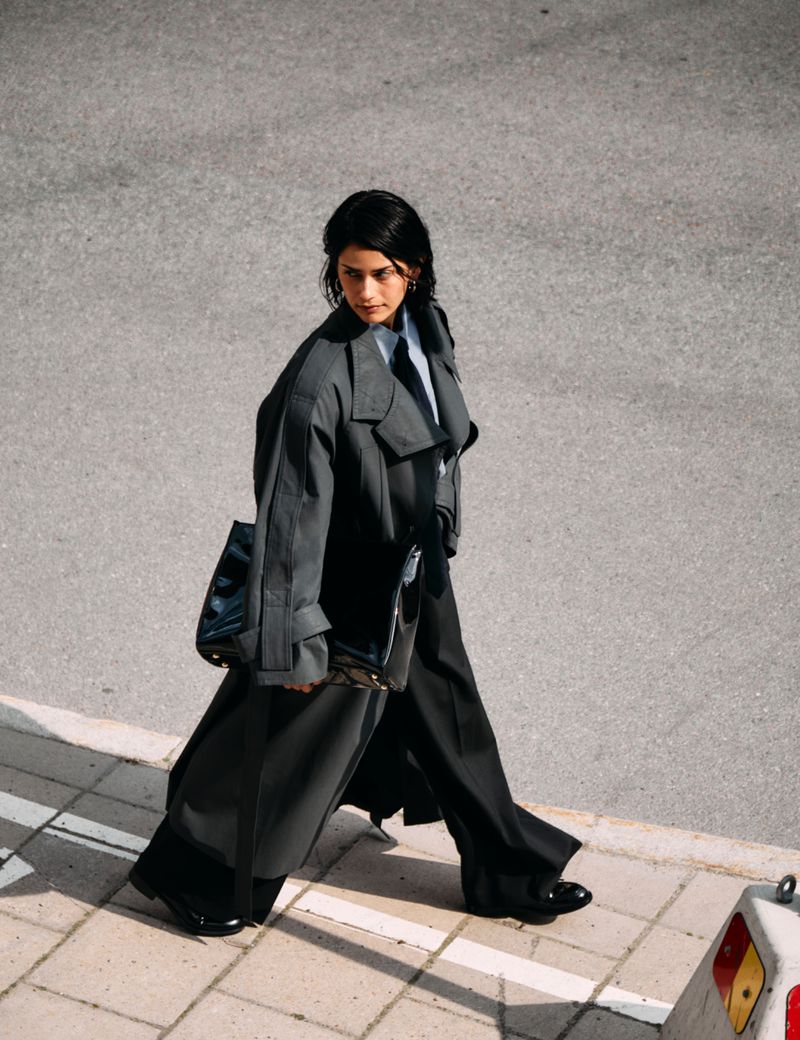 Woman walks on pavement in oversized grey trench coat and black wide-leg pants, carrying a black bag in sunlight. Fashion editorial.