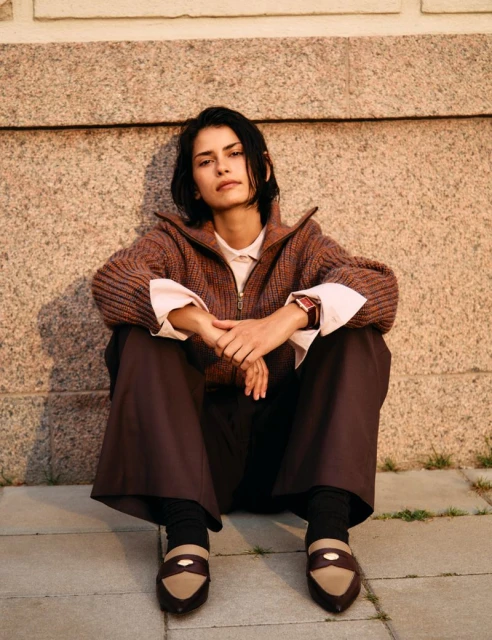 A woman sits on a stone surface wearing a brown cardigan, trousers, socks and beige and brown loafers, gazing directly at the camera.