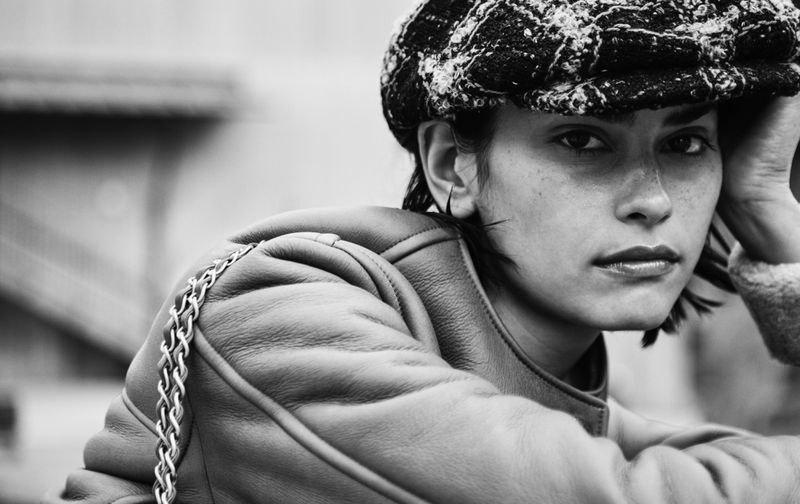 Black and white portrait of a woman wearing a cap and leather jacket with a chain strap, posing in an outdoor setting.