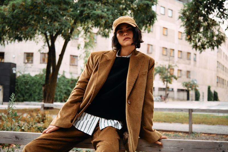 Adult female with a neutral expression poses in a brown corduroy suit with a black shirt and baseball cap in an urban environment.