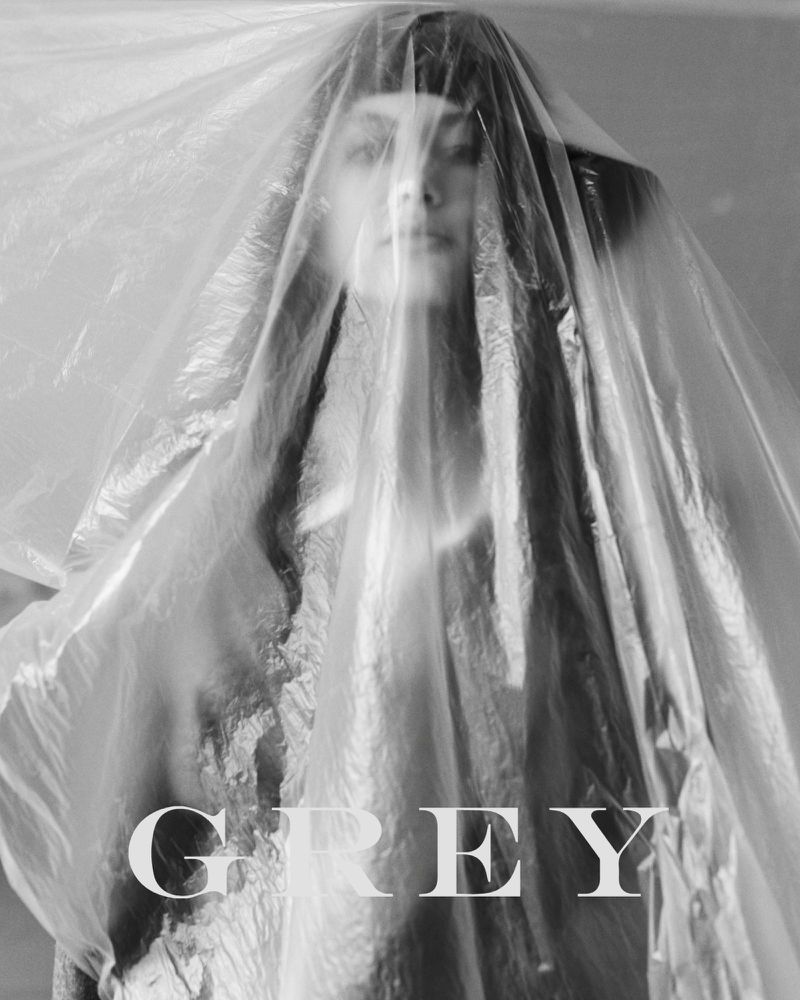 Monochrome portrait of a woman's face partially obscured by a translucent plastic sheet, with the word 'GREY' superimposed.