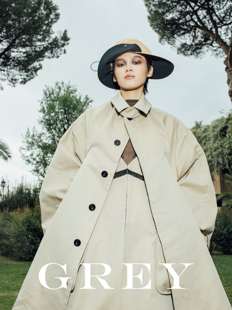 Fashionable woman in a beige coat and wide-brimmed hat poses outdoors in an urban park setting with trees and greenery in the background.