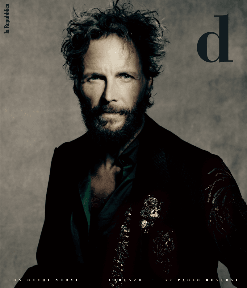 Portrait of a fair-skinned adult male with curly hair and a beard, wearing a decorative jacket, on the cover of a magazine.