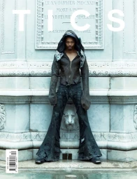 Model standing in front of an ornate stone monument wearing a dark hooded jacket and flared trousers with distressed details.