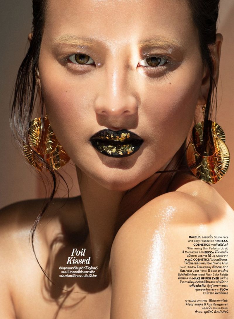 Close-up portrait of an adult female model with gold foil eye makeup and glossy black lipstick with gold foil flakes, plus gold earrings.