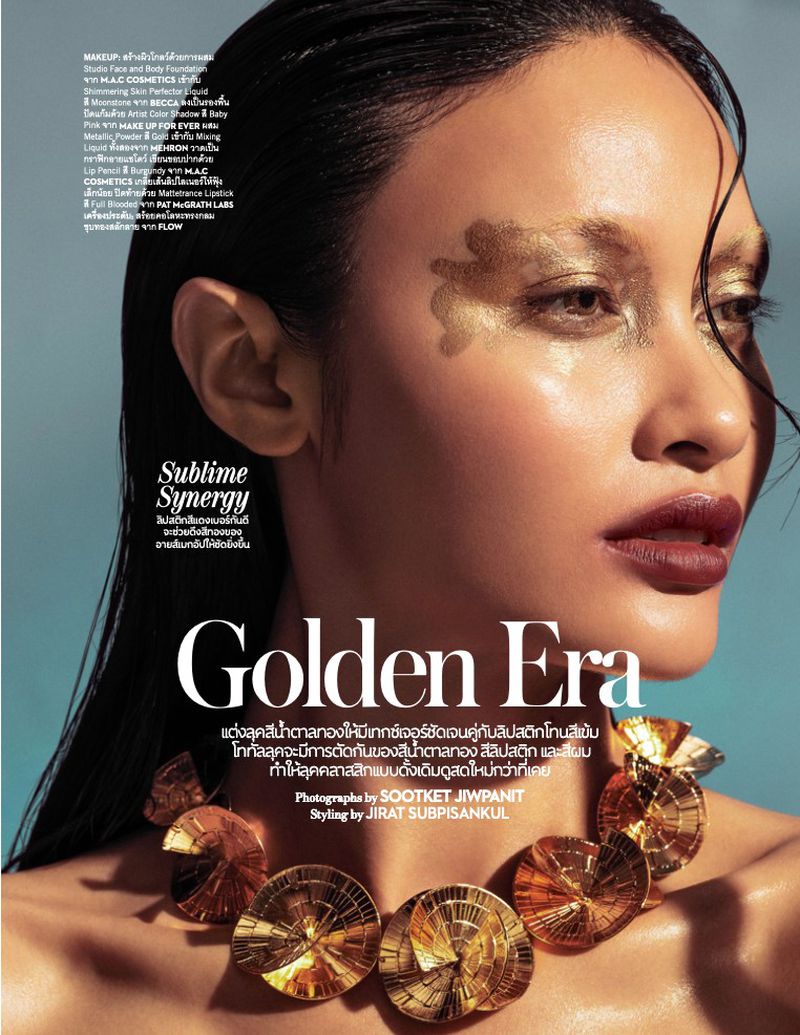 Close-up editorial shot of a woman with golden eye makeup and a gold disc necklace, wearing deep red lipstick against a light blue backdrop.