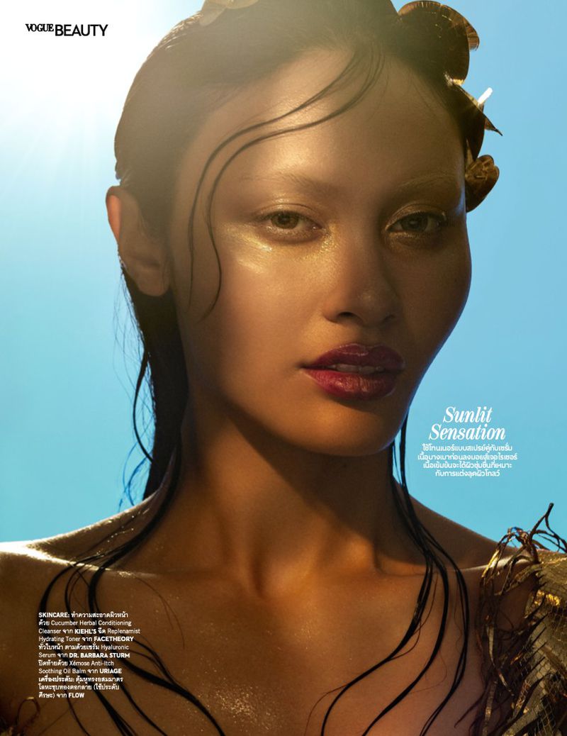 Close-up of an adult female fashion model with tan skin, glossy makeup, and wet-look hair, set against a bright blue background and golden sunlight.