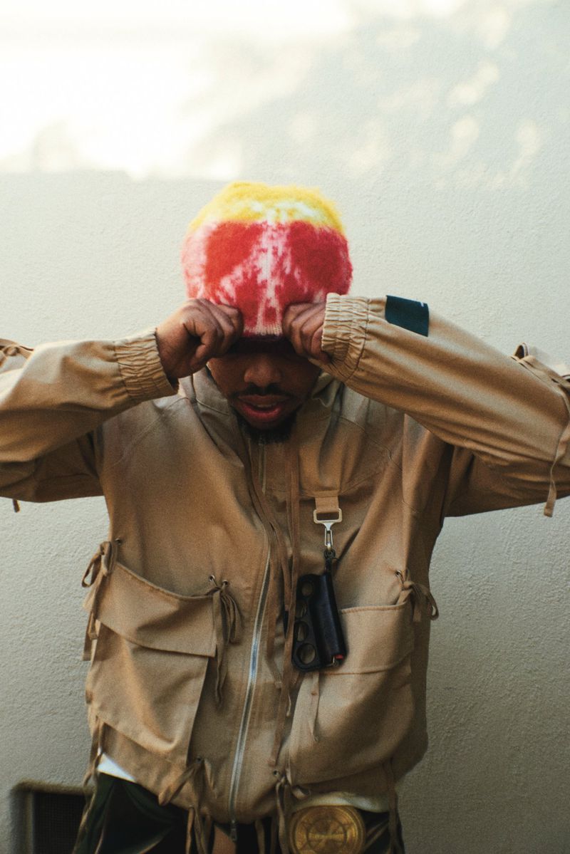 Adult man wearing a colorful beanie and tan jacket with pockets, hands on head, standing against a beige wall with neutral lighting.