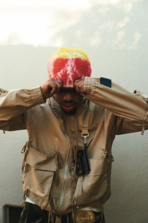 Adult man wearing a colorful beanie and tan jacket with pockets, hands on head, standing against a beige wall with neutral lighting.
