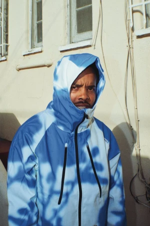 Earl Sweatshirt wearing a blue and white patterned hooded jacket outside against a pale building exterior