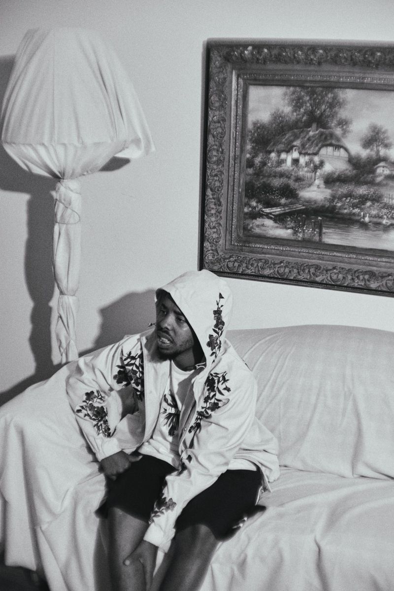 Monochrome photo of an adult male in patterned hoodie sitting on a white couch, with a covered lamp and framed painting in the background.