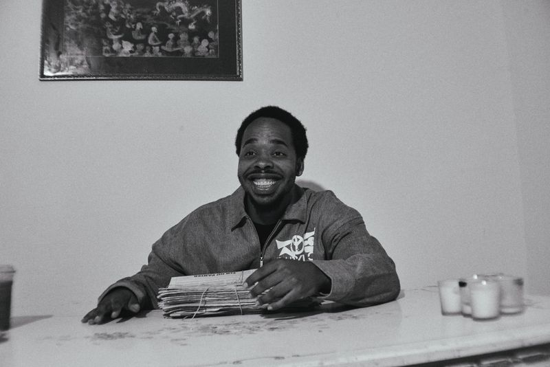 Monochrome medium shot of smiling adult man at table with papers and candles. Simple indoor background with framed picture.