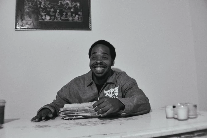 Monochrome medium shot of smiling adult man at table with papers and candles. Simple indoor background with framed picture.