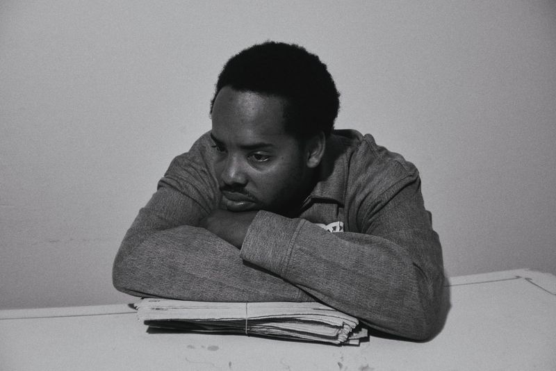 Monochrome portrait of an adult male with dark hair resting his chin on his hands over a stack of newspapers on a white table.