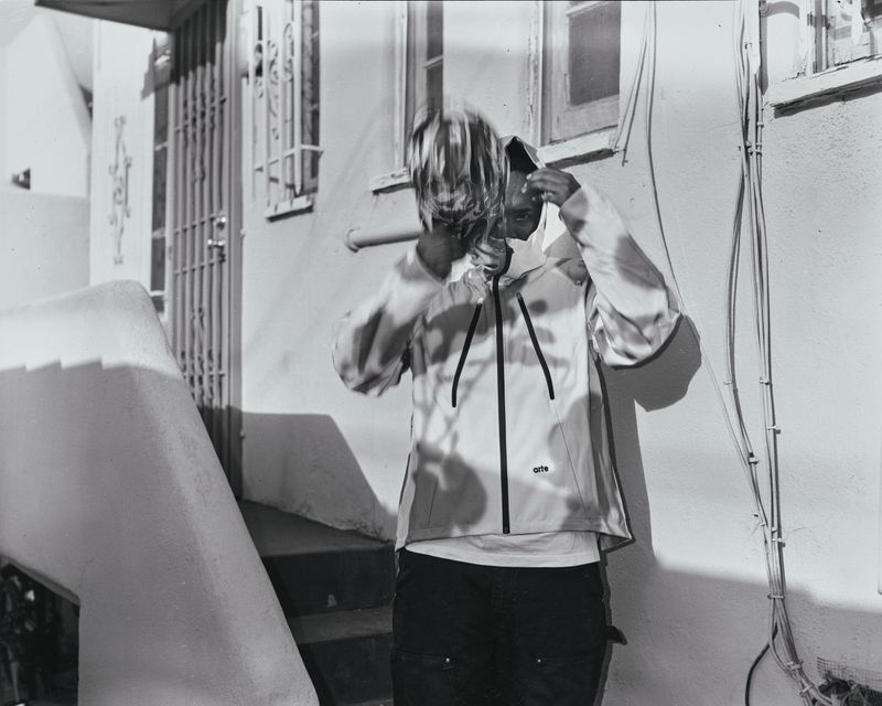 Monochrome shot of a person in a jacket adjusting their hood near a white building with stairs and metal gate, outdoor daylight.