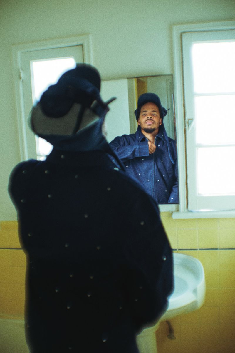 Earl Sweatshirt looking into a mirror in a bathroom wearing a dark jacket