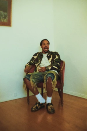 Man with dark skin sitting in red armchair in light-filled room with wood floor wearing patterned jacket and shorts with shoes and white socks.