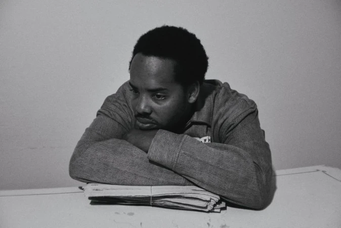 Earl Sweatshirt resting his arms on a tied stack of newspapers against a plain background