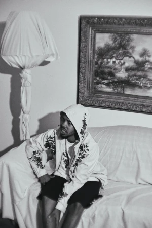 Monochrome portrait of Earl Sweatshirt seated on a draped sofa in a domestic interior with a framed landscape painting and floor lamp.
