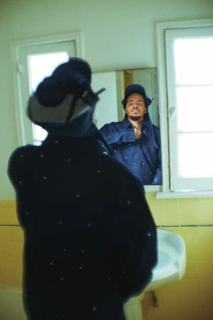 Earl Sweatshirt looking into a mirror in a bathroom wearing a dark jacket