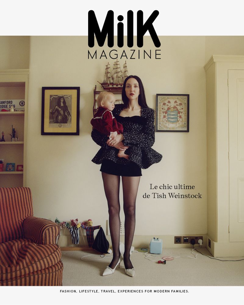 Magazine cover featuring a woman in a patterned jacket and black shorts holding a baby indoors, with a striped chair and artwork.