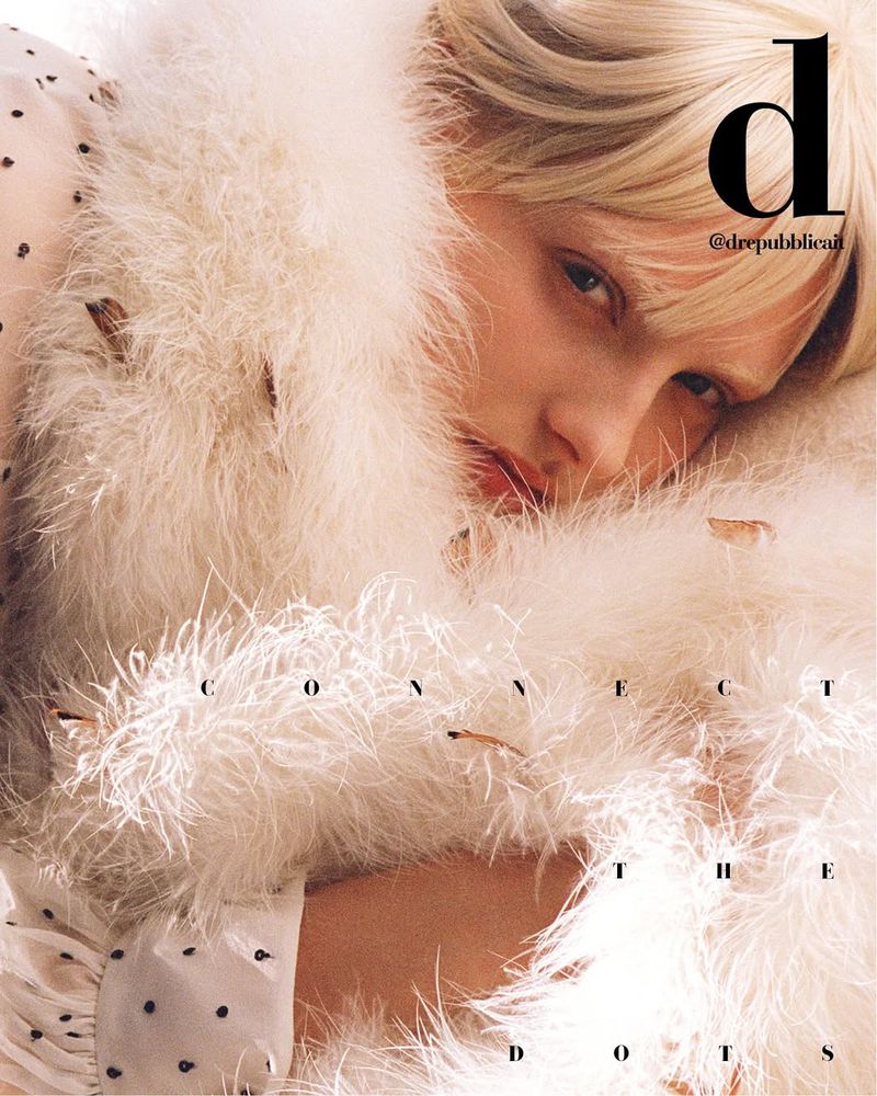 Close-up of a fair-skinned woman with blonde hair surrounded by a feathery boa, looking at the camera with a serious expression.