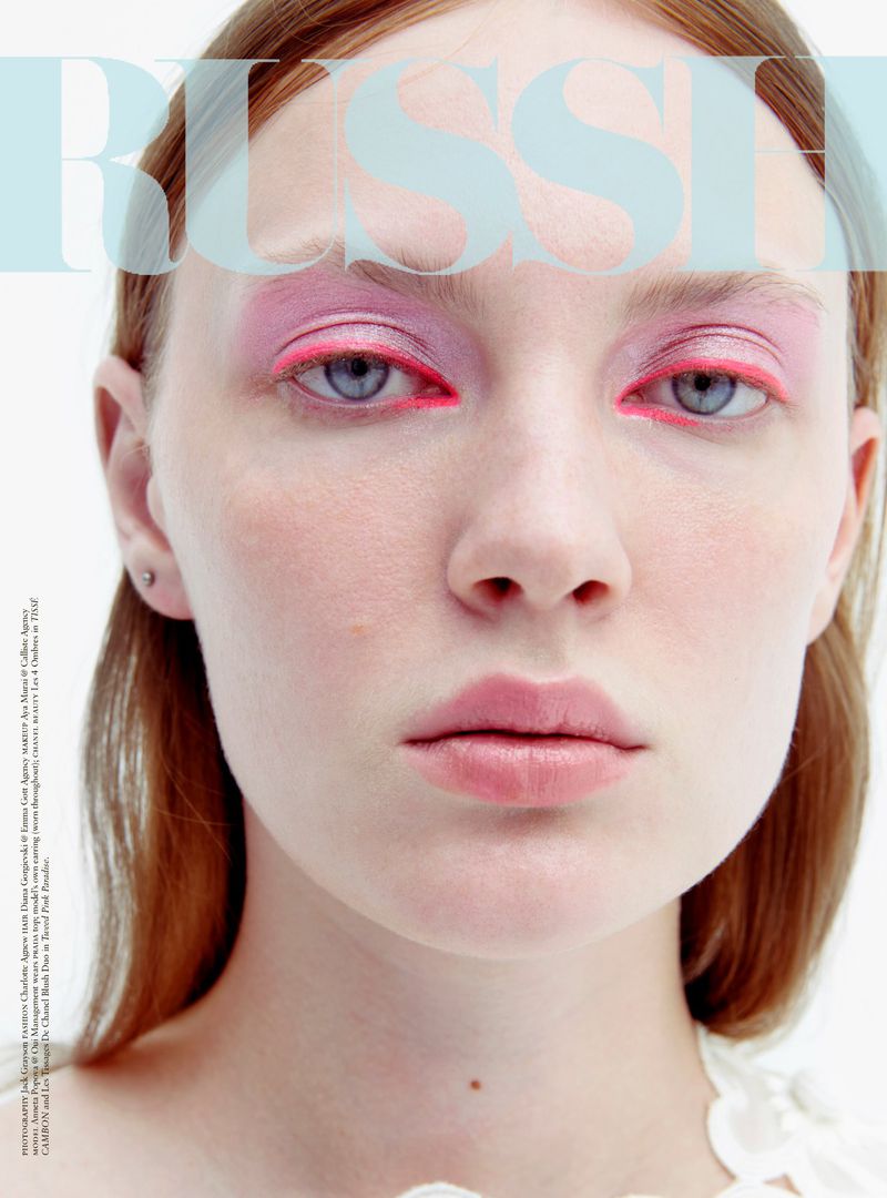 Close-up fashion portrait of a red-haired woman with pink and red eye makeup, fair skin, and light blue eyes in a white studio.
