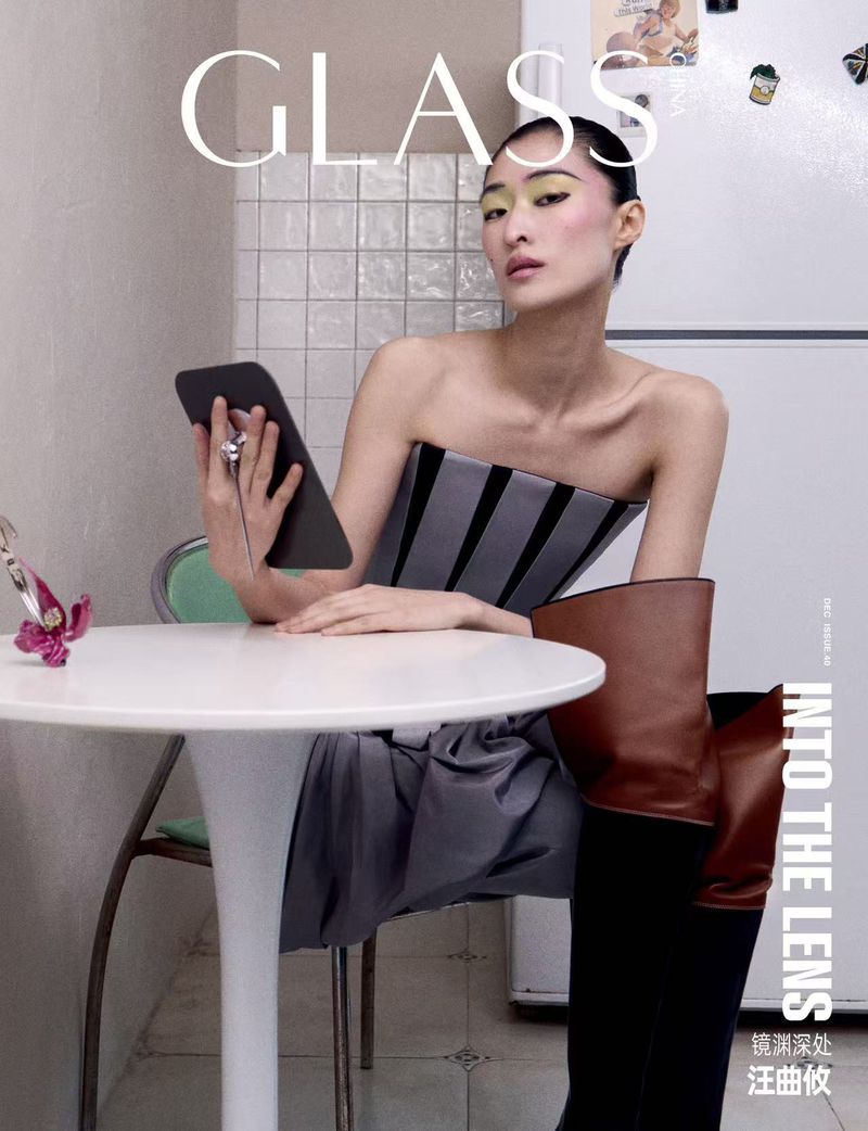 Asian woman with bold makeup and brown over-the-knee boots seated at a table, holding a tablet in a kitchen. Glass magazine cover.