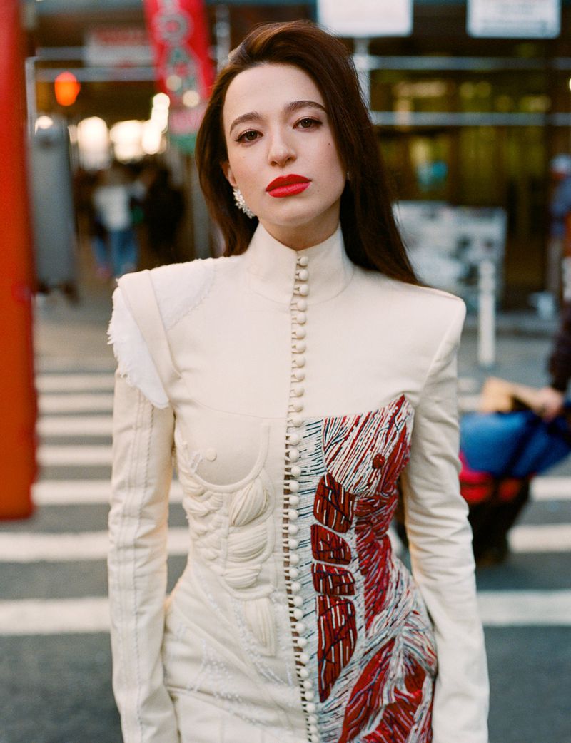 Woman in avant-garde dress with button details poses on city street with red and white architectural details in the background.