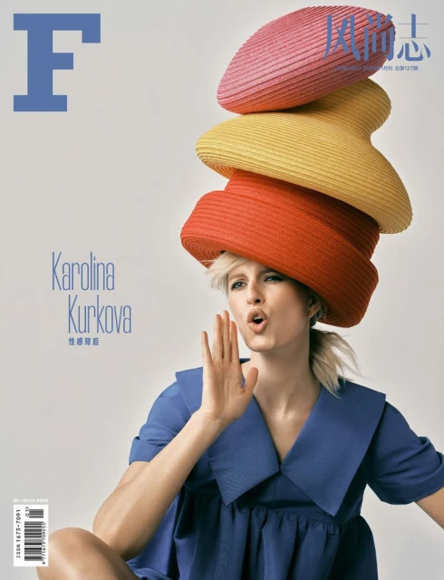 Fashion magazine cover featuring a woman in blue dress and towering hats, posing with hand near her face against a white background.