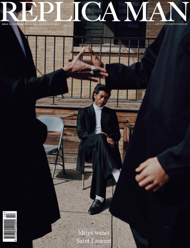 Replica Man magazine cover featuring a seated model in a black suit framed by two other figures against a brick wall backdrop.