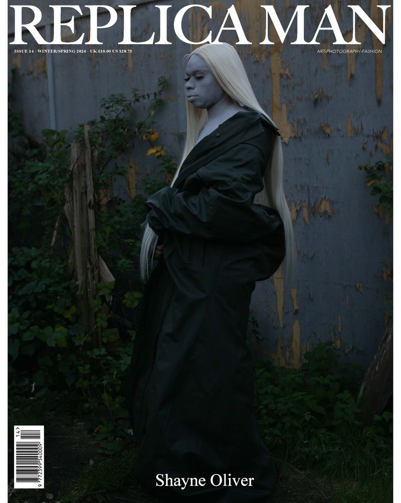 Replica Man magazine cover featuring a model with albino-like skin and long blonde wig, wearing a green oversized trench coat.