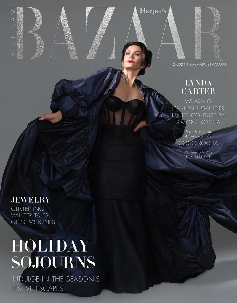 Lynda Carter on Harper's Bazaar Vietnam cover. She wears a black dress and long dark blue coat, posing against a grey background.