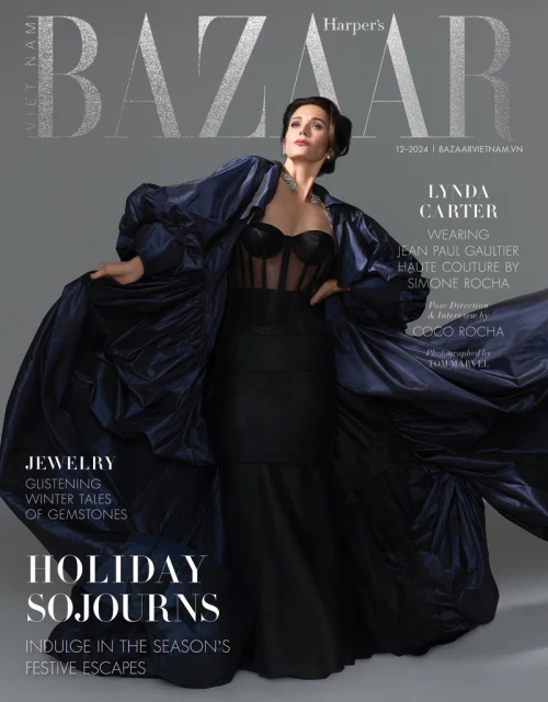 Lynda Carter on Harper's Bazaar Vietnam cover. She wears a black dress and long dark blue coat, posing against a grey background.