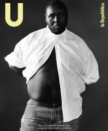 Black man wearing an unbuttoned white shirt and jeans against a dark studio background for U Repubblica magazine