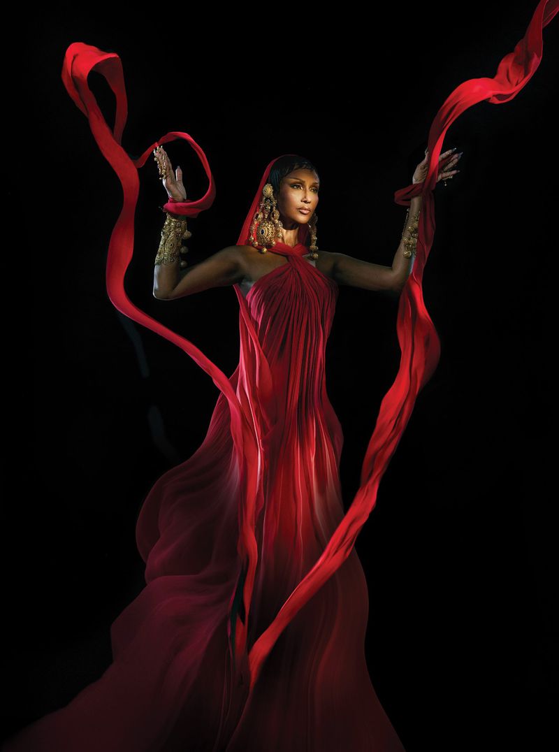 Elegant woman in a flowing red dress and gold jewelry poses in front of a black background, holding flowing red fabric above her.