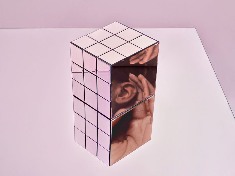 Abstract cube structure with mirrored images of hands and ear against a pale pink backdrop, creating a surreal, dimensional art piece.