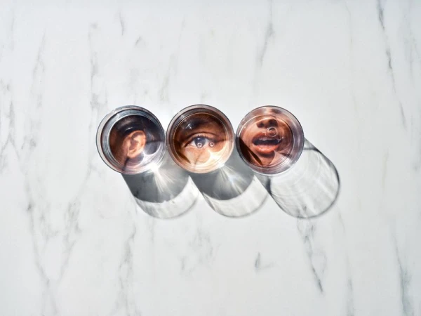 Three glass cups filled with water show refracted images of a human ear, eye, and mouth on a white marble surface.