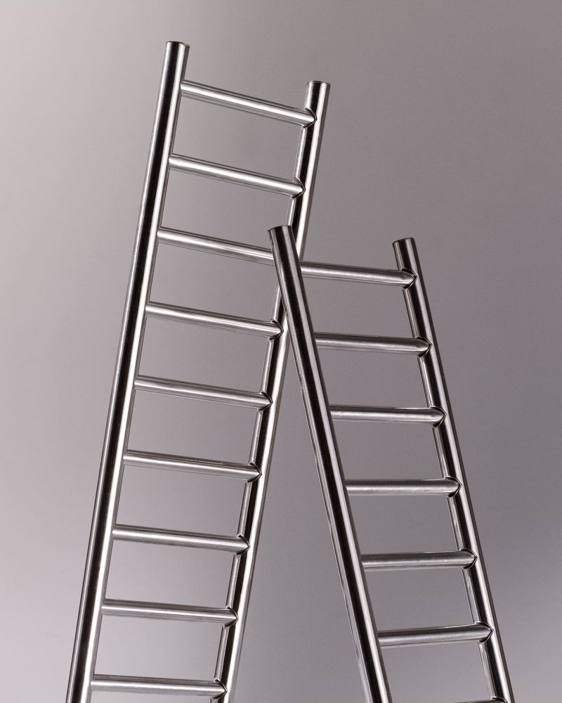 Two shiny metal ladders of different heights lean against a gradient gray background in a studio shot emphasizing their structure.