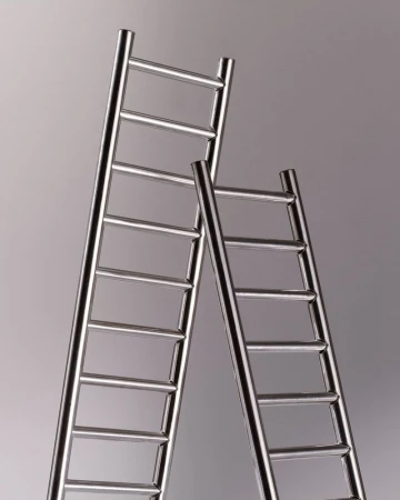 Two shiny metal ladders of different heights lean against a gradient gray background in a studio shot emphasizing their structure.